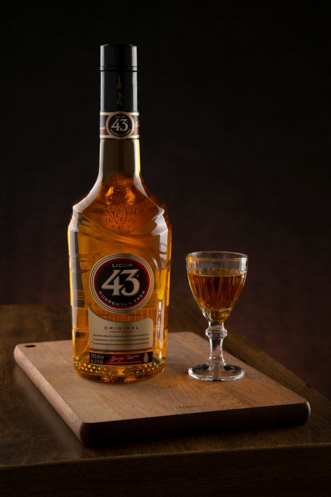 A bottle of Licor 43 and a glass on a wooden board with dark background.